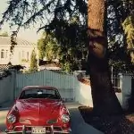 Volkswagen Karmann Ghia rojo vintage estacionado bajo un árbol con un edificio detrás